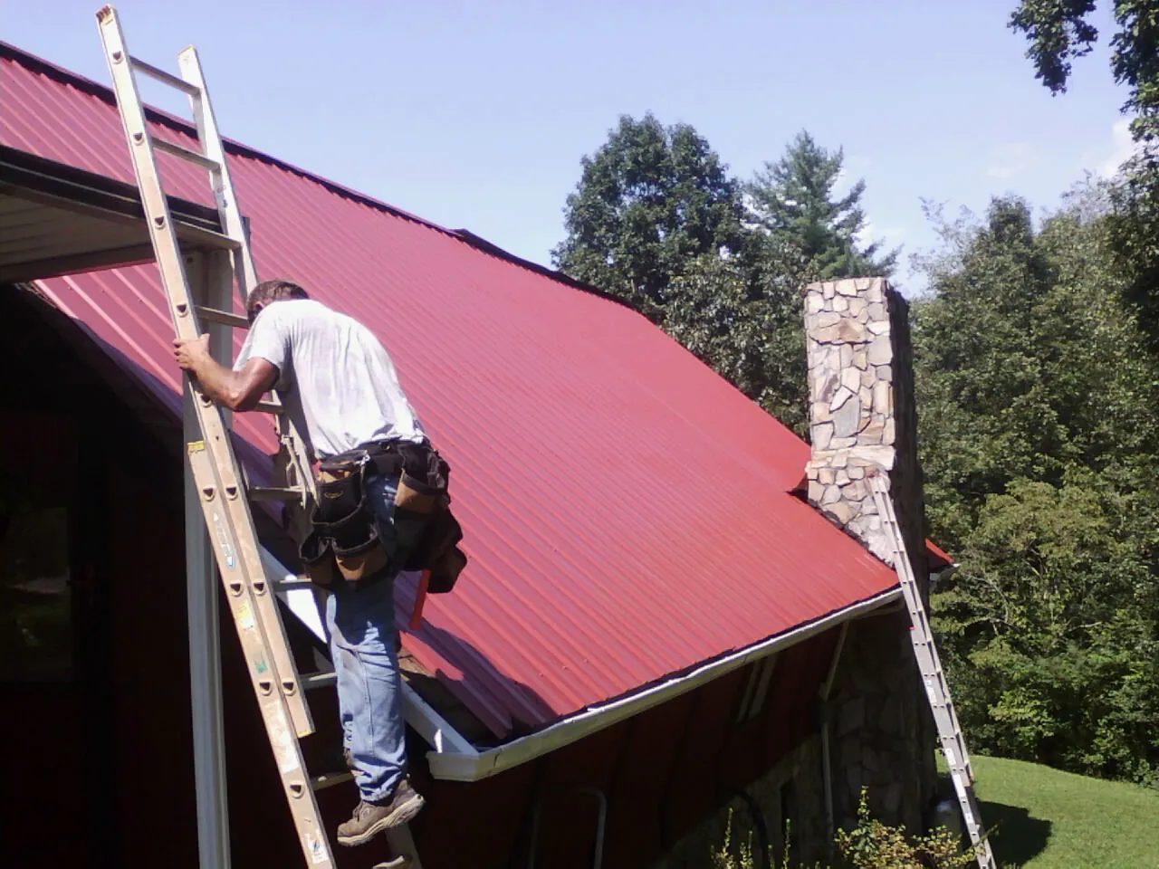 Roofer on a ladder beside a red metal roof and stone chimney — Soffit & Fascia Repair by Roofing Brothers in Prairie City